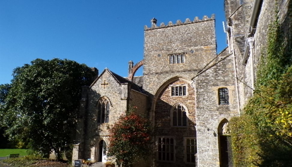Buckland Abbey's south elevation against a blue sky. Green grass and small trees frame the entrance to the building.