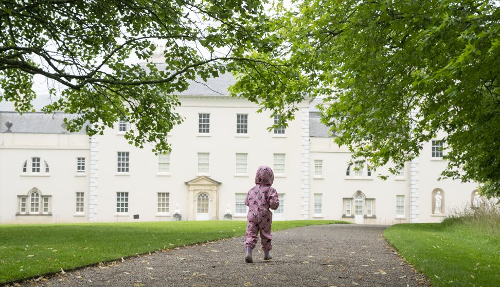 A little girl wearing a pink rainsuit runs towards Saltram House