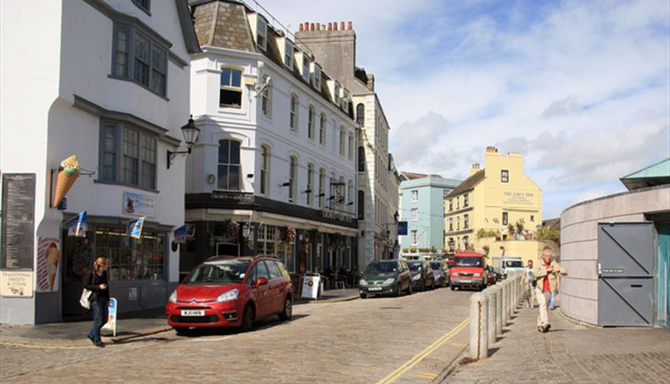 Looking at the outside of the Crown & Anchor on a cloudy day from across the cobbled street which has cars parked on it.