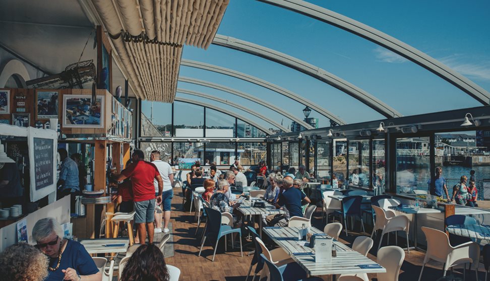 Inside the Boathouse Café with the roof retracted. Customers sitting at tables with a blue sky and views over Sutton Harbour.