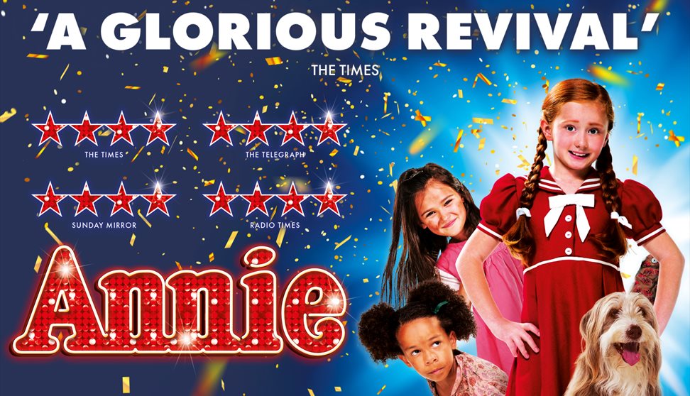 Children and dog stand amongst confetti. Annie is central in a red dress.