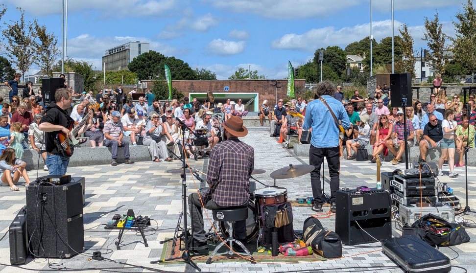 A crowd sits on tiered stone steps around an outdoor plaza amphitheatre, watching performers seated in a circle on the paved ground under a bright blu