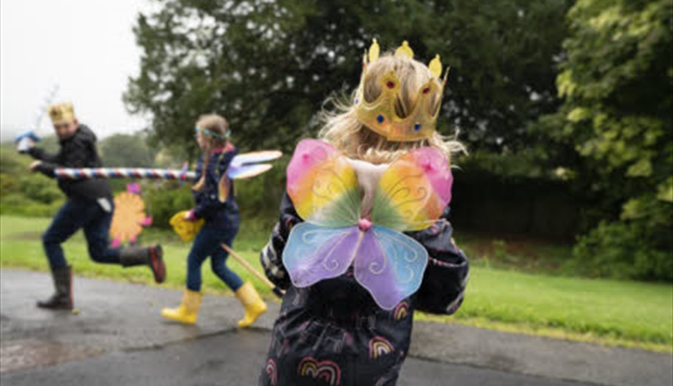 A child with rainbow fairy wings faces away from the camera in the foreground, with other children out of focus in the background.