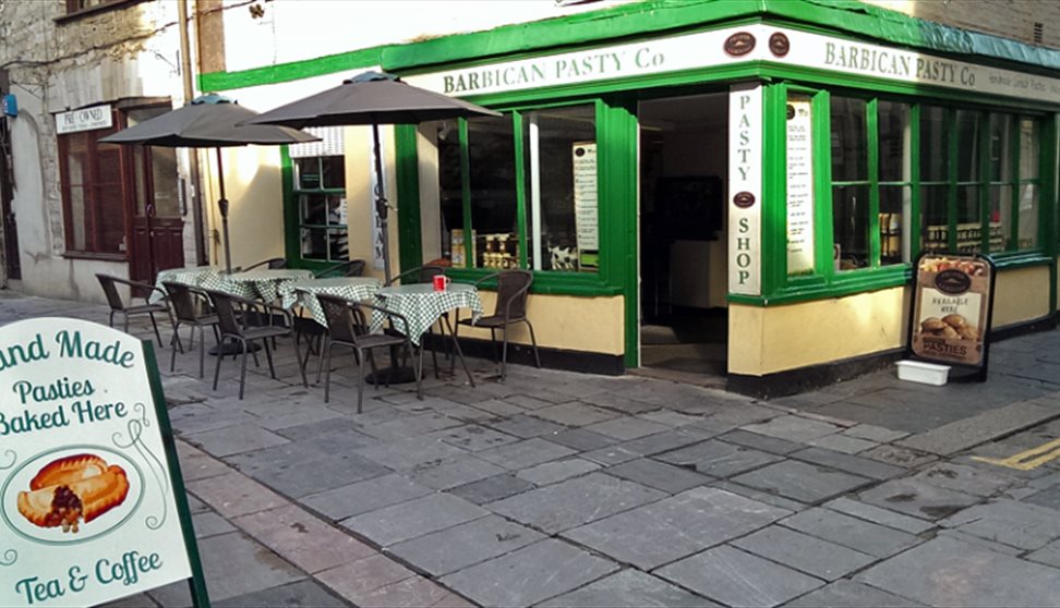 The green, white and yellow painted outside of the Barbican Pasty Co. with outside seating and tables and a sign.