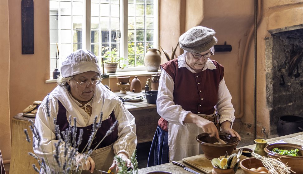 Two women in Tudor costume stand at a wooden table covered in herbs and pottery.