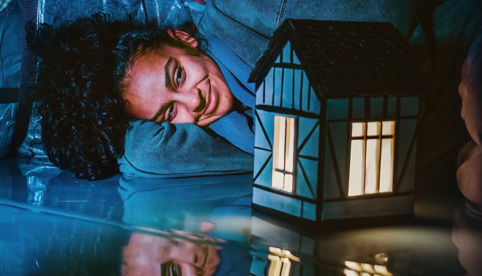 A woman lies with her head on her arm. She smiles, looking at a model of a house which is lit up with a golden light. Everything else is lit up in blu