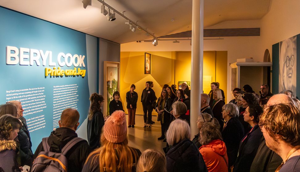 A large group of people listen to a talk in the 'Beryl Cook: Pride and Joy' exhibition at The Box Plymouth