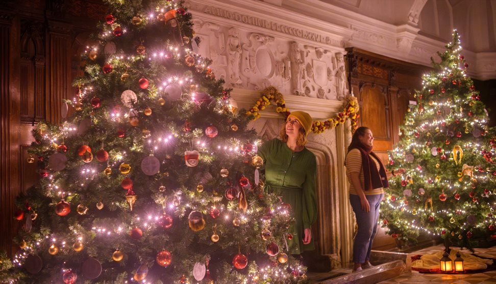 Two large Christmas trees either side of a fireplace, with women placing decorations on the branches.