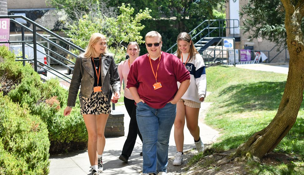 A group of four City College Plymouth students walk together outside on campus on a sunny day. They are smiling and wearing orange lanyards with ID ba