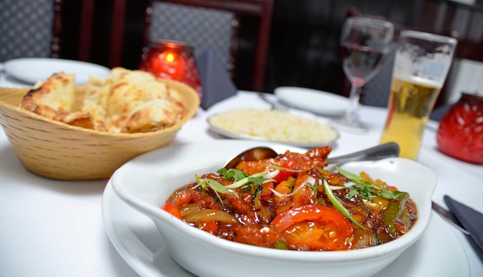 A table set for a meal with a bowl of curry, naan bread in a basket, a plate of rice, a drink and candles.