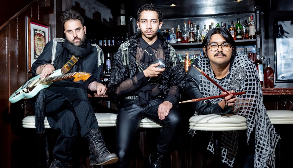 Three men sit in front of a bar. Each hold a different thing: A guitar, a microphone and drumsticks. They wear black with silver embellishments and do