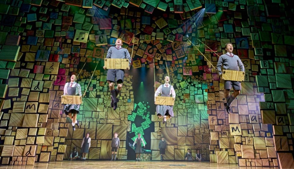 Stage scene from Matilda the Musical with schoolchildren on swings in front of a colourful wall of alphabet blocks.
