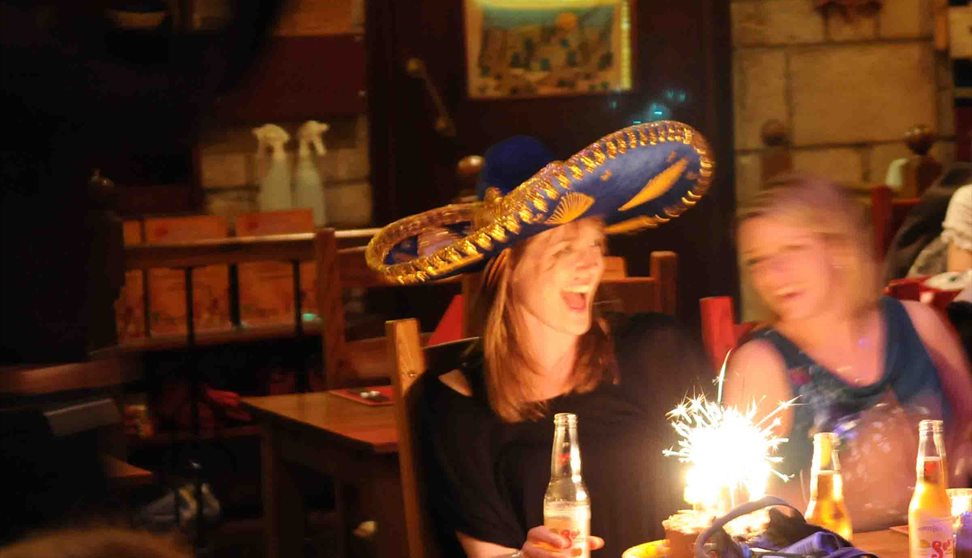 Diners sat at a table with a birthday cake with lit sparkler and a lady smiling and wearing a sombrero.