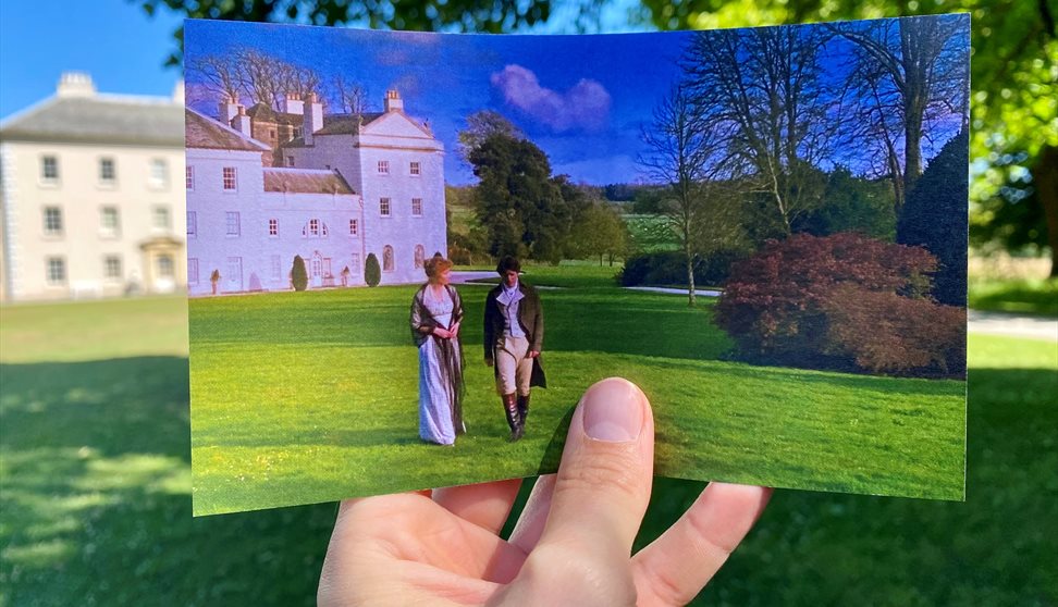 A hand holds a photograph showing two people in historical dress in front of a white estate, matching the real-life scene behind it.