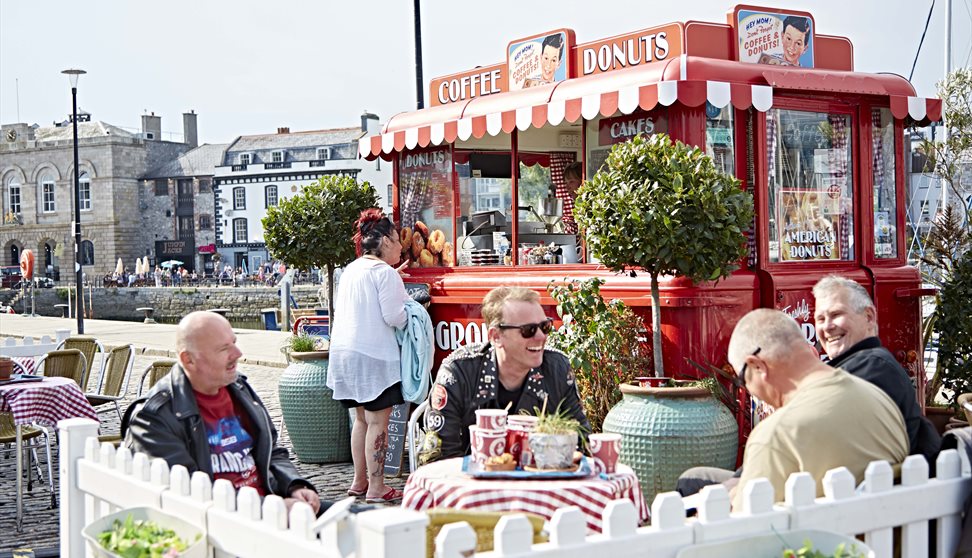 Red vintage Coffee and Donuts van with striped awning, outdoor seating area with Potted trees, white picket fence and customers sat at tables.