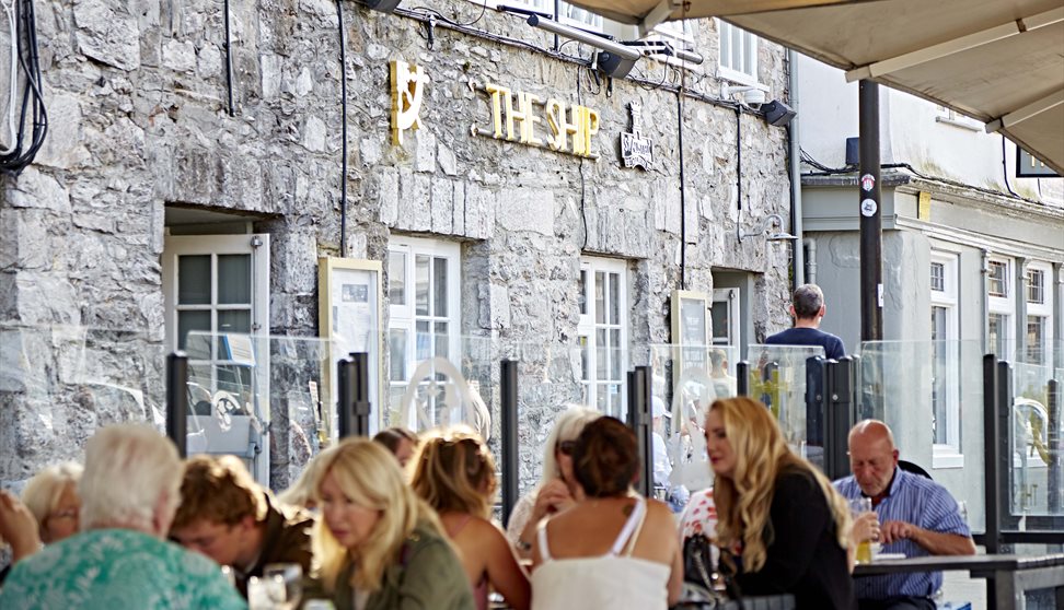 Customers sat at outside seating and tables in front of The Ship. Glass partitions separate the tables and there are large parasols overhead.