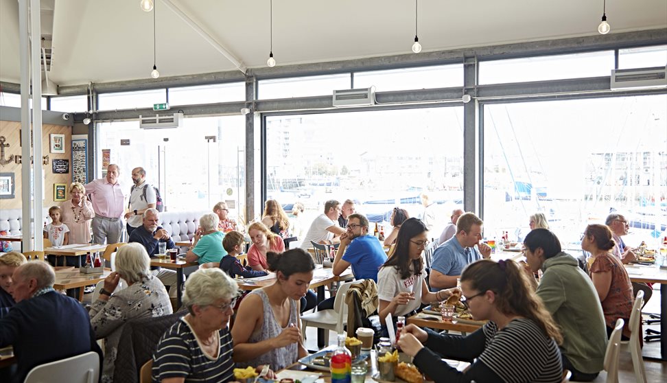 Busy inside of the restaurant showing tables full of diners in front of glass walls with views over Sutton Harbour.