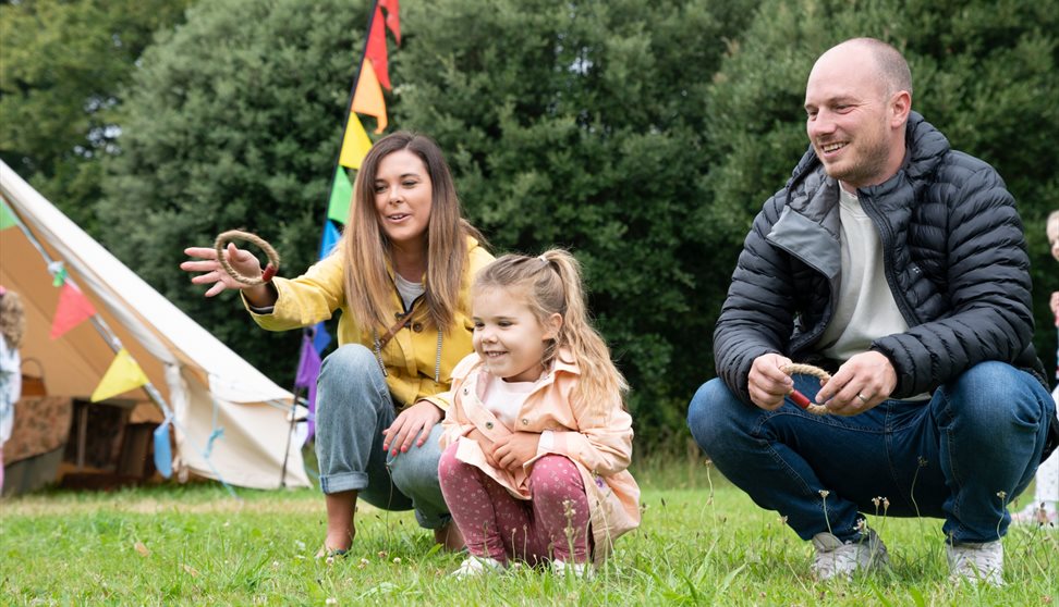 A family of three plays ring toss on grass near a tent with colourful flags; the mother and father kneel while the daughter watches.