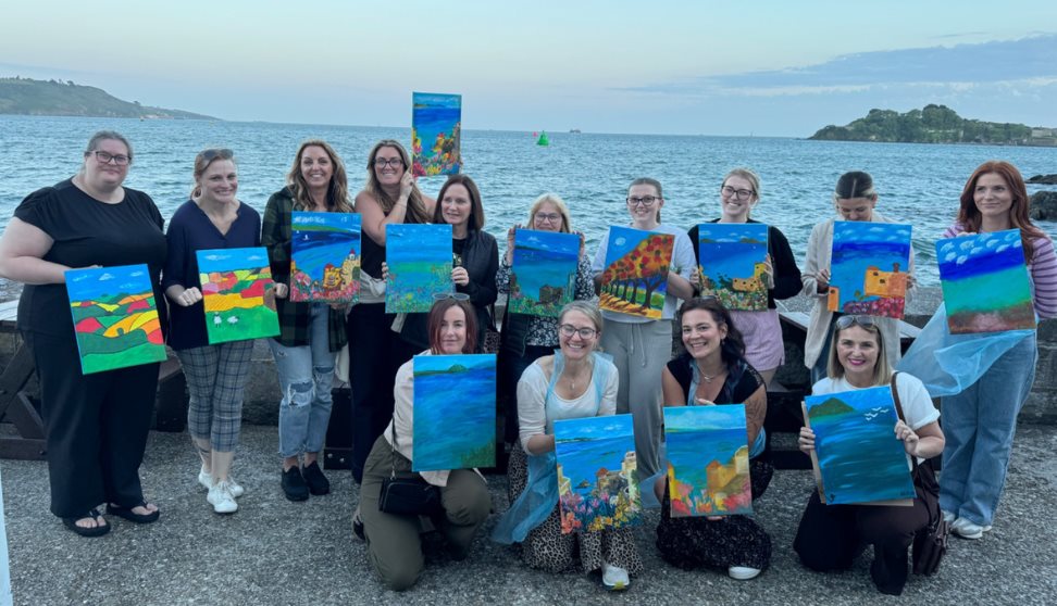 Group enjoying a Sip and Paint evening at The Waterfront in Plymouth, holding their finished paintings by the sea.