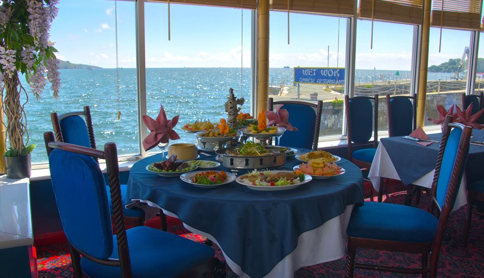 A table inside the restaurant set for a meal with dishes of food in front of large glass windows overlooking the water of Plymouth Sound.