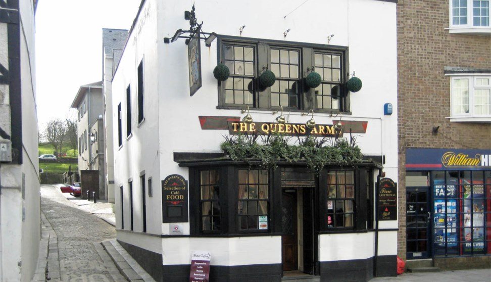 The white and black painted front of the Queens Arms with plant displays above the entrance.