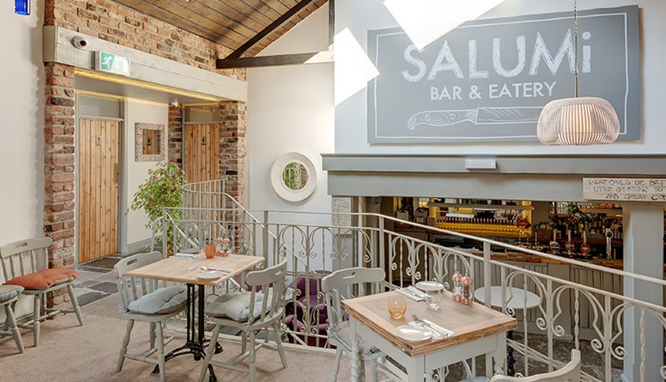 Inside Salumi, showing cream painted walls, tables and chairs, exposed brickwork and cream wrought iron railings separating seating areas.