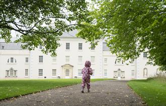 A little girl wearing a pink rainsuit runs towards Saltram House