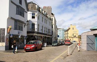 Looking at the outside of the Crown & Anchor on a cloudy day from across the cobbled street which has cars parked on it.