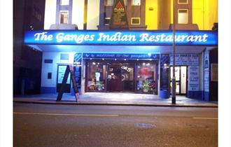 Front of the restaurant showing a brightly lit blue sign saying The Ganges Indian Restaurant.