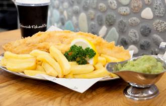 A plate of fish and chips garnished with lemon and parsley and a jug of mushy peas.