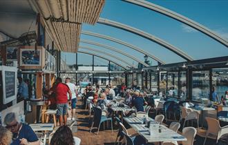 Inside the Boathouse Café with the roof retracted. Customers sitting at tables with a blue sky and views over Sutton Harbour.