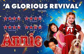 Children and dog stand amongst confetti. Annie is central in a red dress.