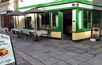 The green, white and yellow painted outside of the Barbican Pasty Co. with outside seating and tables and a sign.