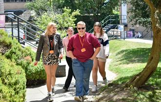 A group of four City College Plymouth students walk together outside on campus on a sunny day. They are smiling and wearing orange lanyards with ID ba