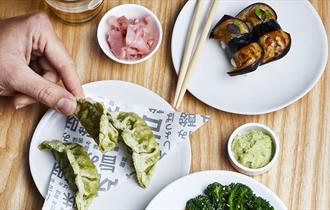 Small white plates of different foods displayed on a table with chopsticks and a hand holding a green gyoza.