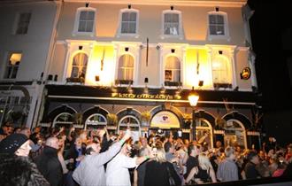 A crowd of people outside the brightly lit front of Kitty O'Hanlons at night-time.