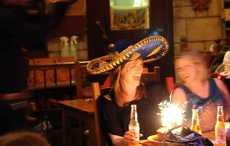 Diners sat at a table with a birthday cake with lit sparkler and a lady smiling and wearing a sombrero.