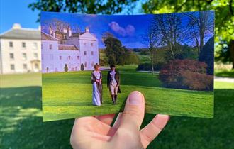 A hand holds a photograph showing two people in historical dress in front of a white estate, matching the real-life scene behind it.