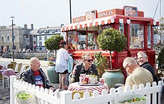 Red vintage Coffee and Donuts van with striped awning, outdoor seating area with Potted trees, white picket fence and customers sat at tables.