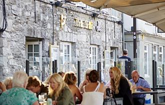 Customers sat at outside seating and tables in front of The Ship. Glass partitions separate the tables and there are large parasols overhead.