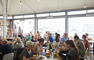 Busy inside of the restaurant showing tables full of diners in front of glass walls with views over Sutton Harbour.
