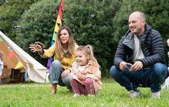 A family of three plays ring toss on grass near a tent with colourful flags; the mother and father kneel while the daughter watches.