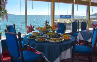 A table inside the restaurant set for a meal with dishes of food in front of large glass windows overlooking the water of Plymouth Sound.