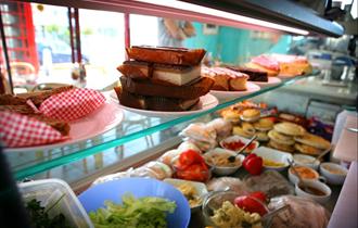 Large refrigerated display cabinet inside shop window displaying cakes, slices, pies and sandwich ingredients.