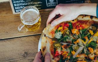 A large pizza divided into portions being pulled apart by a pair of hands. On the table is also a glass of beer and a menu.