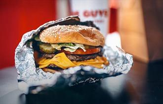 An opened silver foil package containing a large sesame seed topped bun containing a burger with salad and cheese.