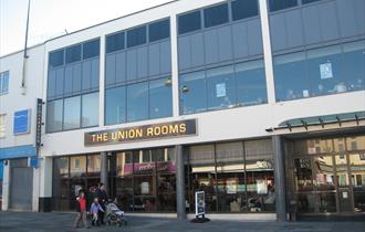 The glass frontage of the Union Rooms on a sunny day with a family and pushchair strolling by in the foreground.