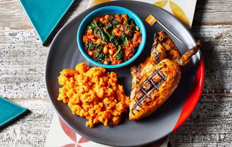 A grey plate with a portion of chicken, sweet potato mash and a green pot of beans and spinach.