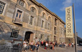 An outside shot of the Seco Lounge against a blue sky showing customers sat at tables and chairs.