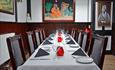 A long table set for dinner with 10 chairs and place settings, coloured folded napkins and red candles.